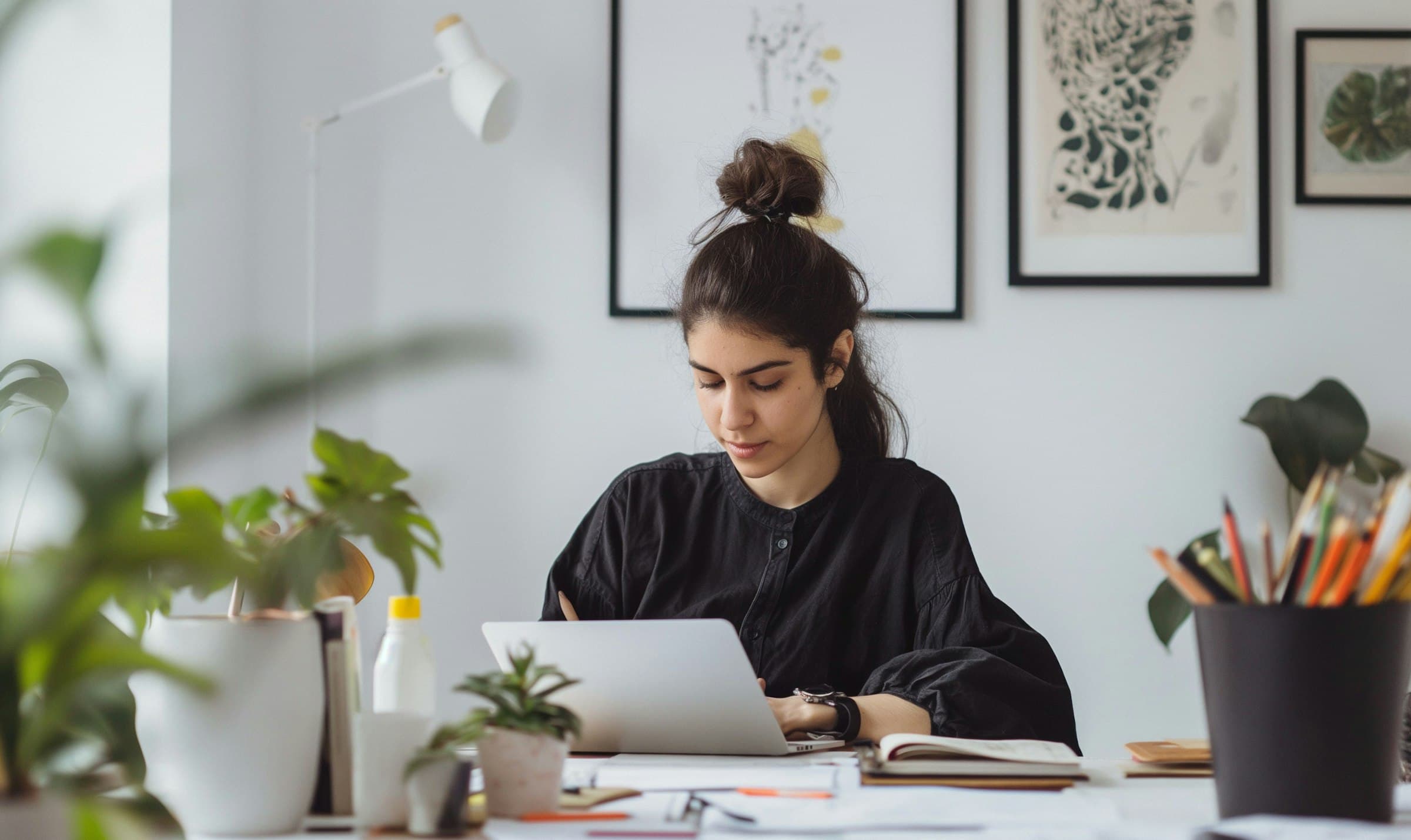Woman working on a laptop at a desk, surrounded by plants and art on the walls, in a bright, modern home office.