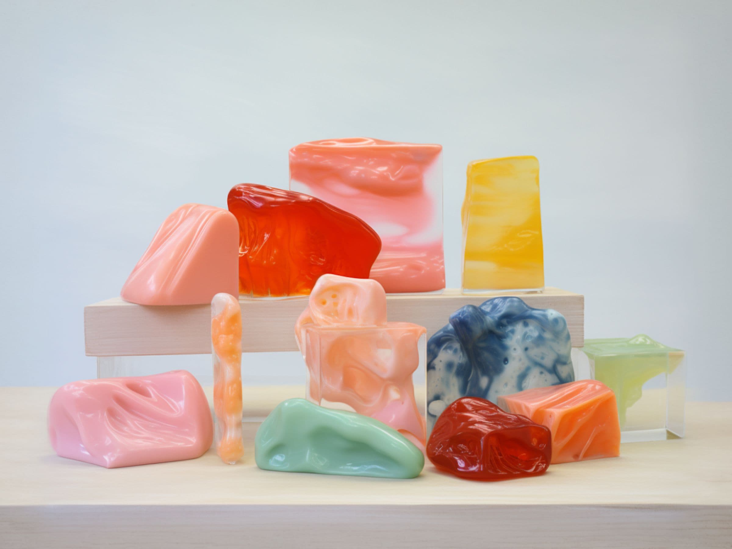 A collection of colorful, abstractly shaped soap bars in pink, red, orange, blue, and green, displayed on a wooden surface and platforms.